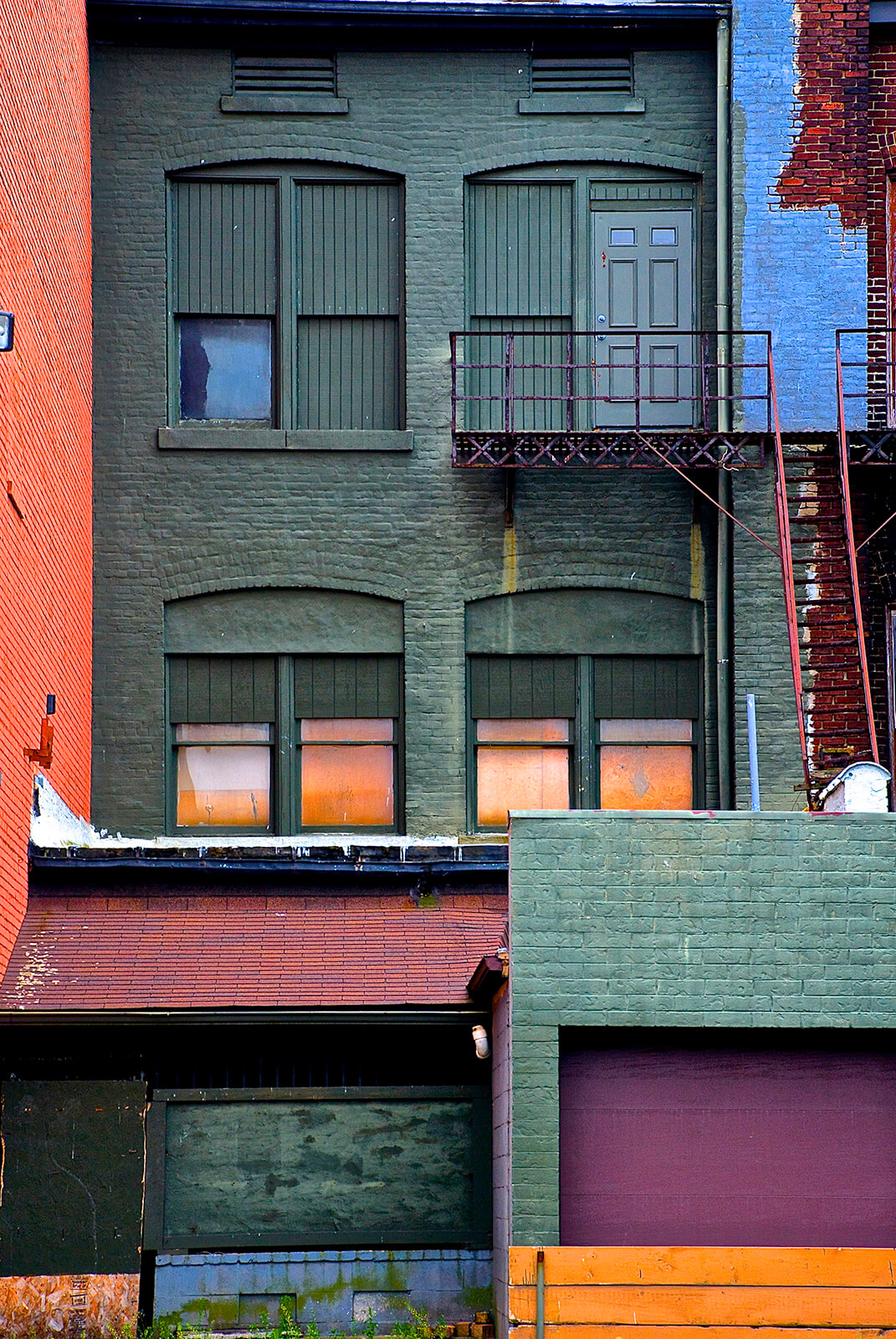 Photograph of a brick building with at least three stories. The image focuses in on a small portion of the building, where you can see the second and third floors, and some of the ground level. The building is painted mostly in a deep forest green, but there are pops of blue-grey, peachy orange, and maroon as well. There is an old iron staircase on the right side that has a rust red coloring and you can see bright orange curtains in the windows of the second floor home.