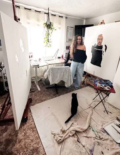An artist stands smiling at the viewer in an indoor art studio. She has large canvases to her left and to her right. The canvas to the right of the viewer shows the early stages of a portrait of a person wearing a black shirt. Behind Autumn is a curtained window and a desk and chair. In front of her, a cat peers up at her while sitting on a protective fabric to catch paint splatters.