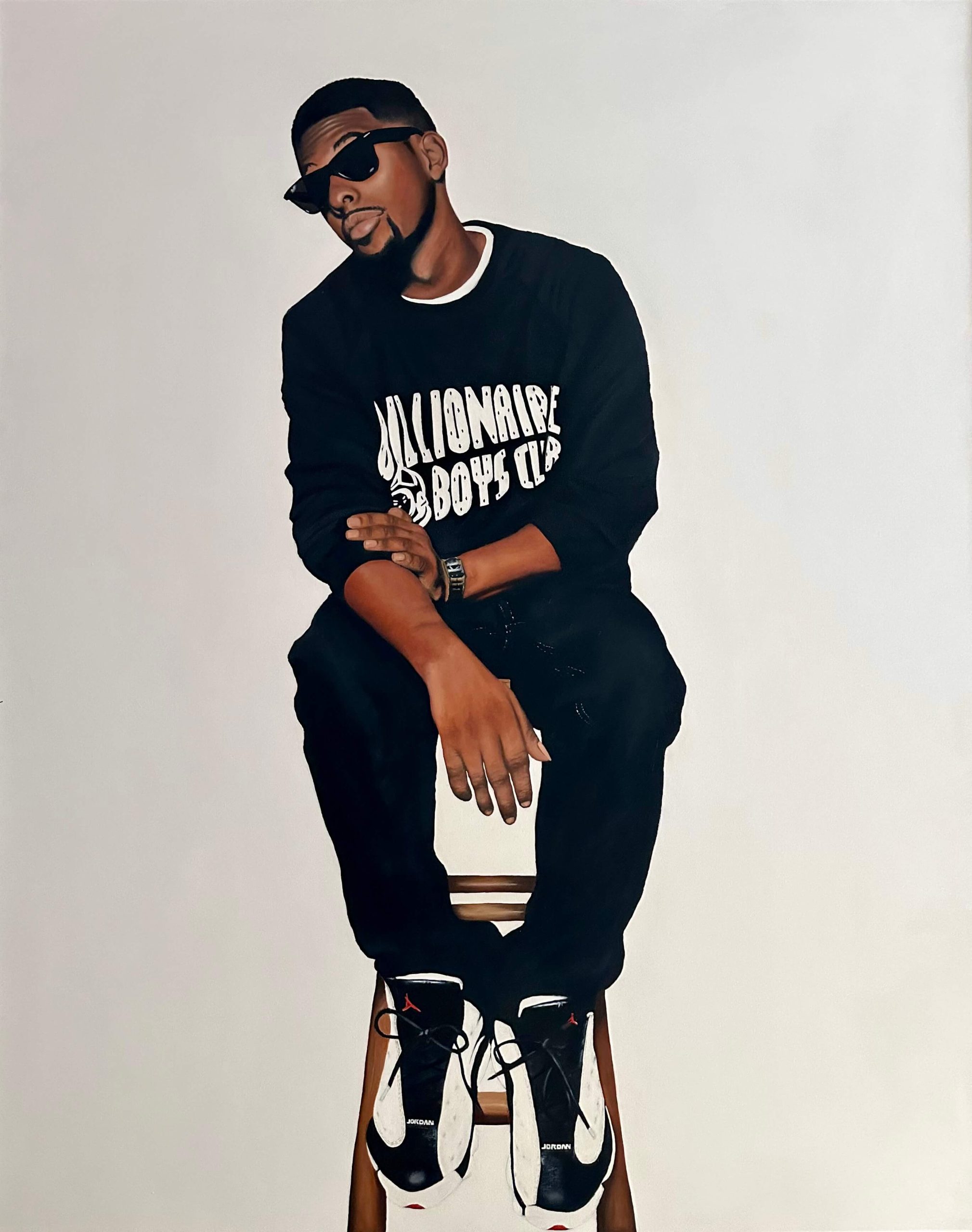 A man sits on a stool; both feet are propped against the lowest rung of the stool, showing his white and black sneakers. He is wearing black sunglasses but still appears to look towards the viewer with his eyebrows raised. The man is young, Black, has short cropped hair, and wears a black long sleeved t shirt with the sleeves rolled up to his elbows and black jeans. His arms are resting on his knees, one hand gently grasping onto the other elbow.