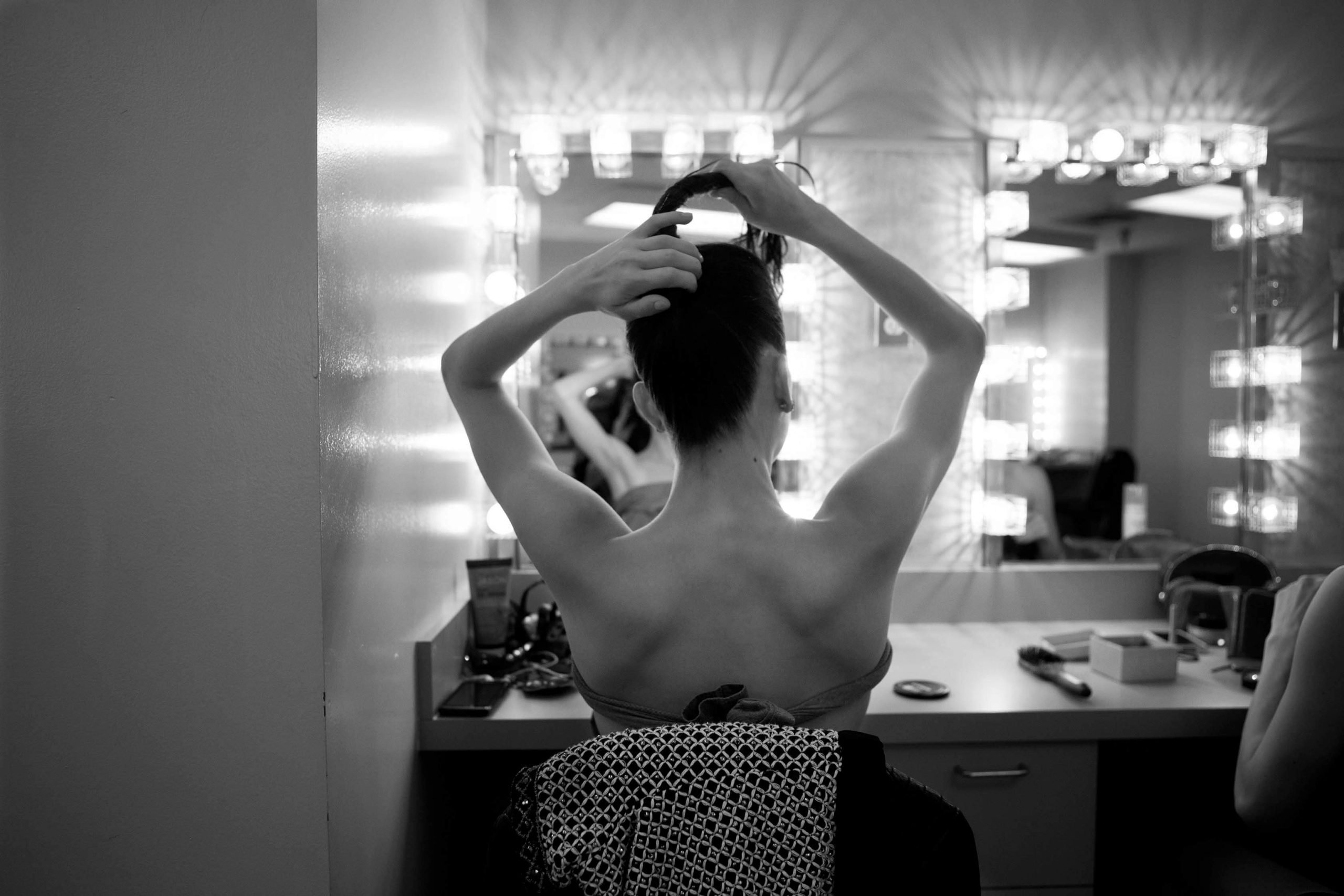 A black and white photograph of a ballerina backstage in the dressing room, sitting in front of a large mirror with bright lights above the mirror. Her arms are up in the air, fixing her hair pinned up. Her shoulder blades are exposed.
