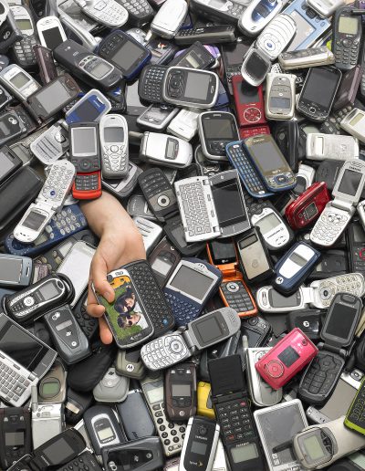 Photo of a bunch of cell phones from the early 2000's in a pile with an arm sticking out holding a cell phone