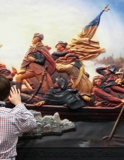 A large tactile display of George Washington and a group of Revolutionary Soldiers crossing a river. A blind man is touching the display with both of his hands.