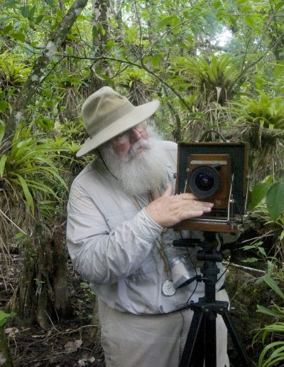 A photo of an older caucasian man with a mustache and long white beard wearing a hat, adjusting an early 20th century camera in the forrest