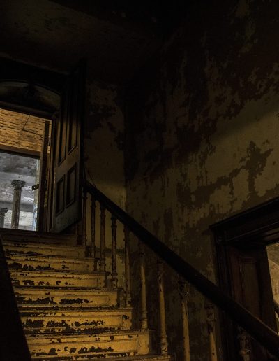This is a photo of a staircase in an old building. Paint is peeling off the walls and stairs. There are railings on both sides of the stairs and there's an open door at the top of the staircase where there are 2 visible pillars.