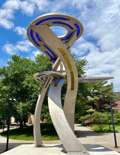 Three swerving rectangles going vertical from a square platform. The rectangles are holding up a circular shape with the center cut out. In the circle are cutouts that have blue stained glass. In the rectangles there are cutouts that have yellow stained glass.