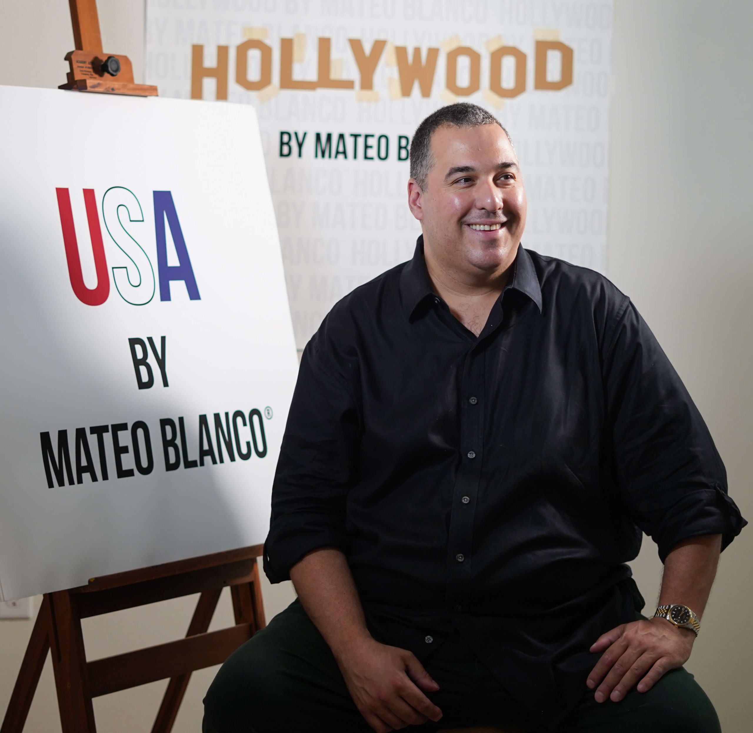 A photograph of artist Mateo Blanco sitting on a chair in front of an easel that displays a sign that reads "USA by Mateo Blanco. In the background is an image with wording that reads "Hollywood by Mateo Blanco."