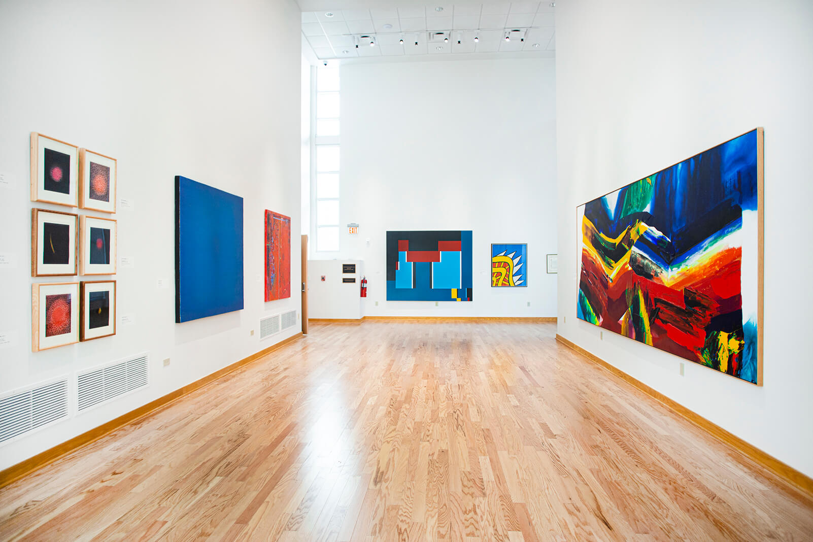 Image of an art gallery with art on the walls on either side, and at the other side of the room. The art on the right is one very large canvas with bold hues of blue, red, yellow, green, and white. On the left are several smaller canvases aligned neatly beside and above each other. The far wall has a combination of large and small art pieces with bold vibrant colors and shapes.