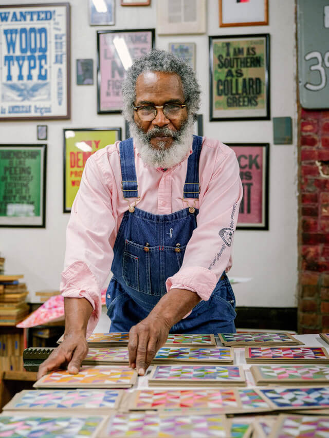 A medium-brown skinned man with salt & pepper hair and a full beard wears overalls and a pink button up shirt. He stands behind a table with stacks of geometric works.