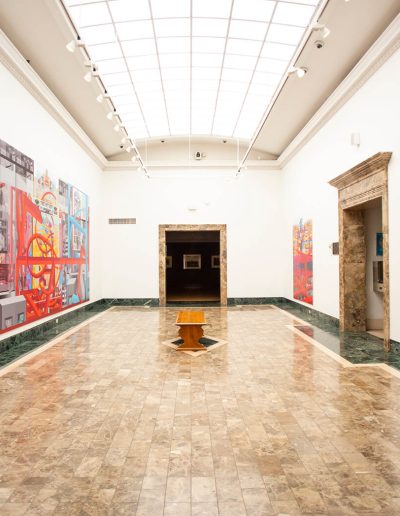 View of gallery with colorful paintings along the left and right walls. A wooden bench sits in the center of the tiled floor.