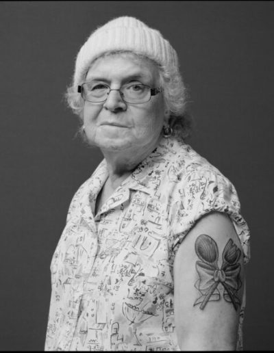 A photo of an older woman with a hat and patterned shirt wearing glasses that has a tattoo of cooking utensils wrapped in a ribbon