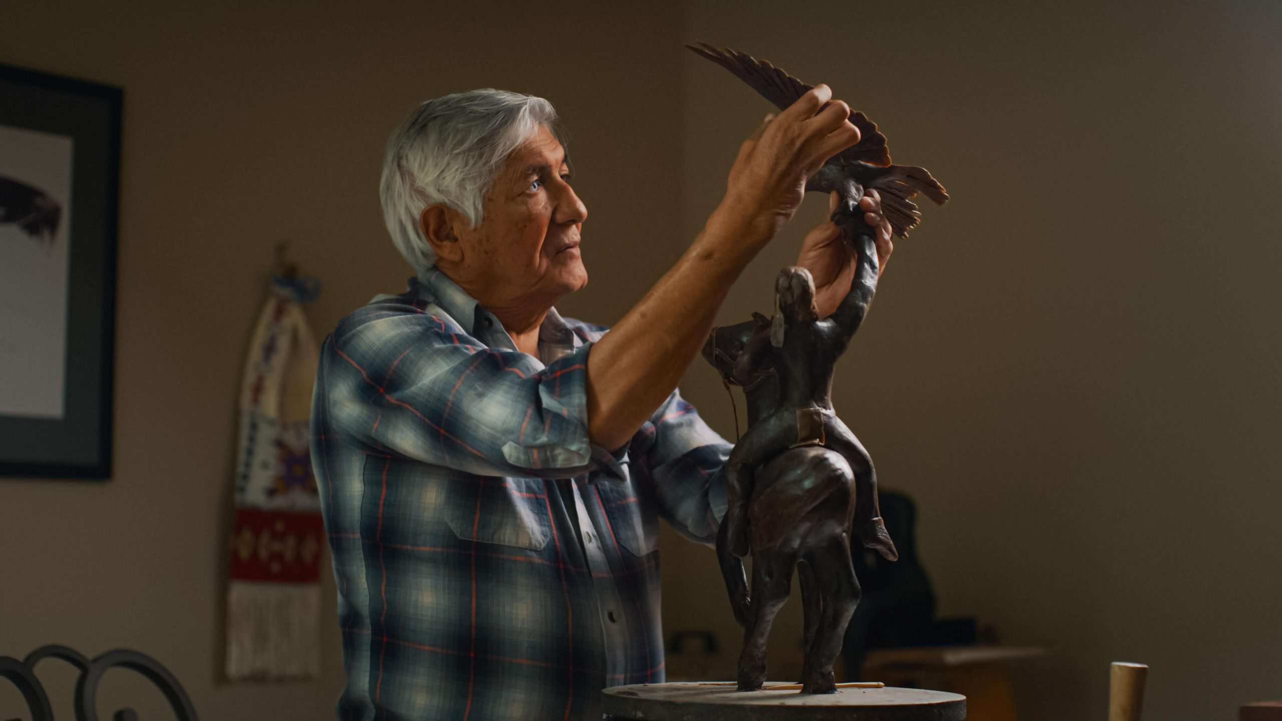Photo of the blind Native American artist Michael Naranjo working on a sculpture. His hands are touching the top of the sculpture, which is a man riding a horse and holding an eagle with its wings spread to fly.
