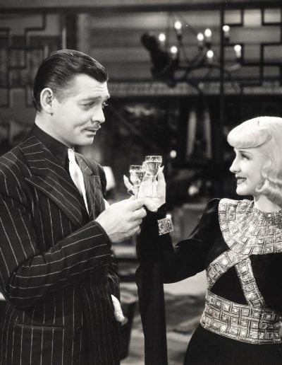 Black and white old style photo of a man and woman clinking champagne glasses. The man is wearing a black suit with white pinstripes and the woman is wearing a formal dress
