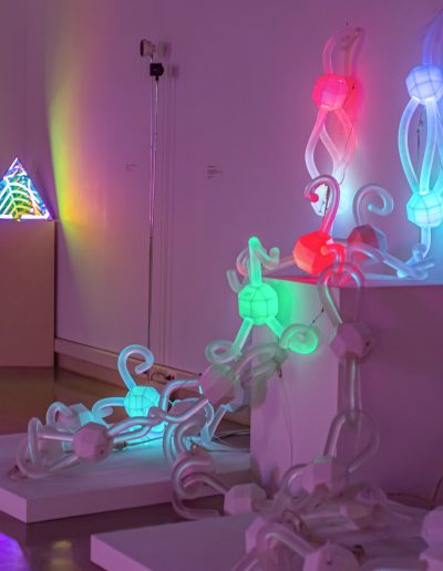 a glass sculpture made of chain links with built-in red, blue, and green LEDs. The lights reflect and blend to create shifting patterns of color and shadow on nearby walls, forming a dynamic, abstract experience through light.