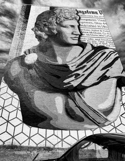 Black and white image. It depicts a bust of Caesar painted over top of a news article and various geometric patterns. It appears to be painted on a large brick wall outside. The cloudy sky is visible behind it.