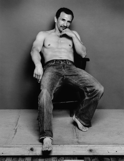 Black and white photo of Ray Mancini sitting in a chair shirtless, wearing jeans and no shoes with his left hand on his chin