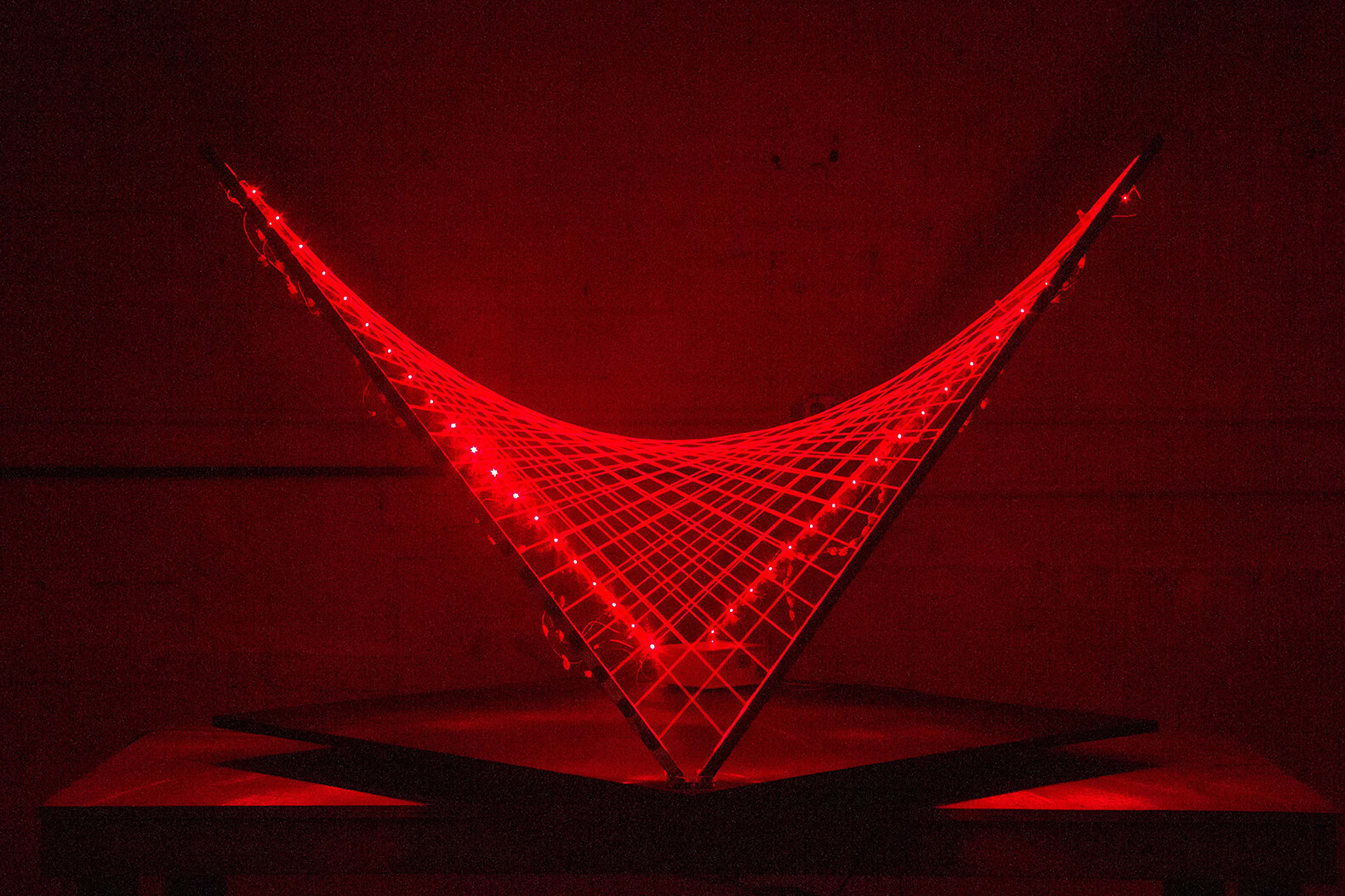 Photo of a warped square with two opposite points touching the stage and the other opposite points bending upward. The warped square acts as a frame that houses multiple red LED lights that shine a fence like webbing across the inside of the frame.