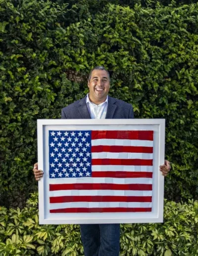 Image of artist, Mateo Blanco, standing in front a green wall of shrubbery. He is wearing a navy blue suit, and is holding one of his art pieces, which is an American flag, made of what appears to be strips of paper, and held together in a white frame. The artist has a wide grin on his face, almost as though he is laughing.